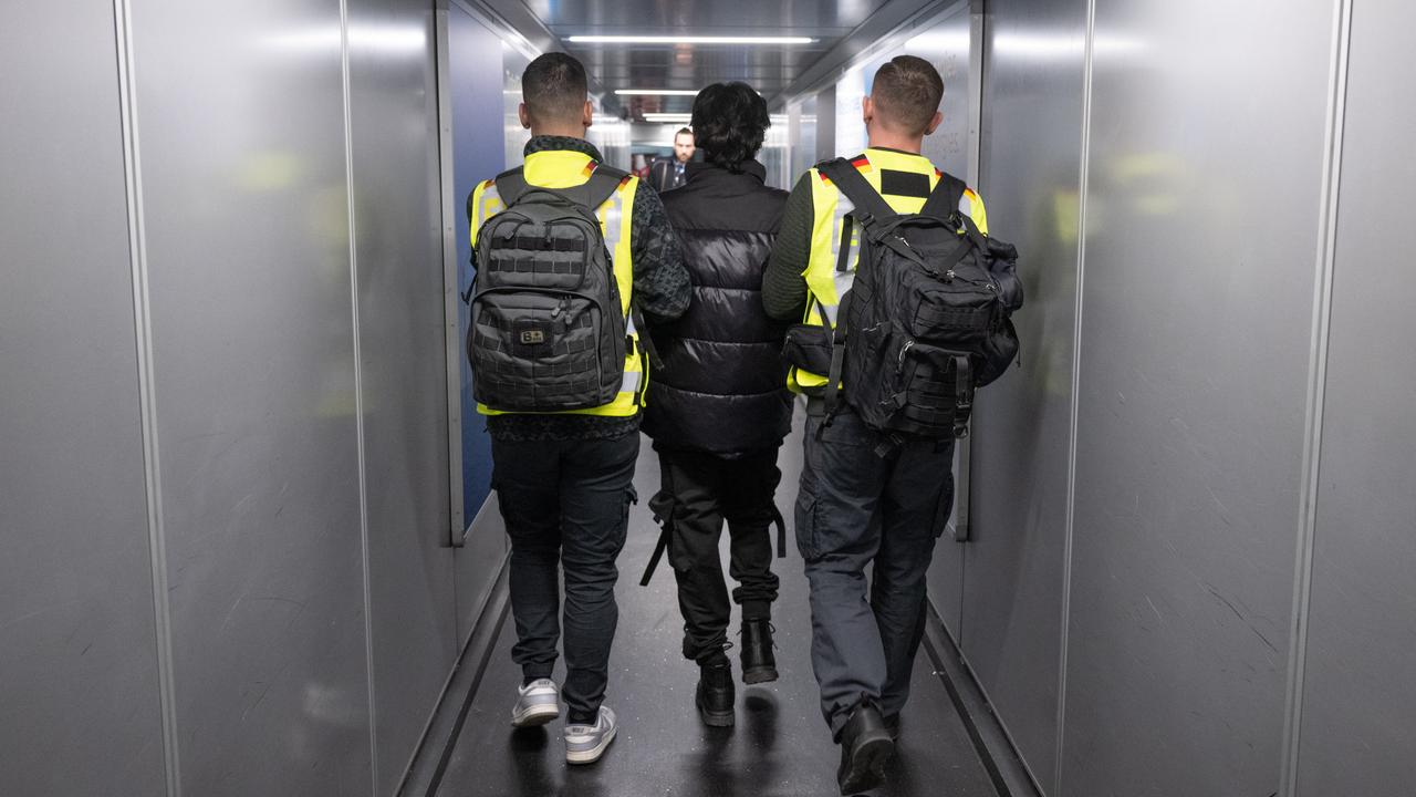 Zwei Beamte in Warnwesten eskortieren einen Mann durch einen Flugzeugtunnel.