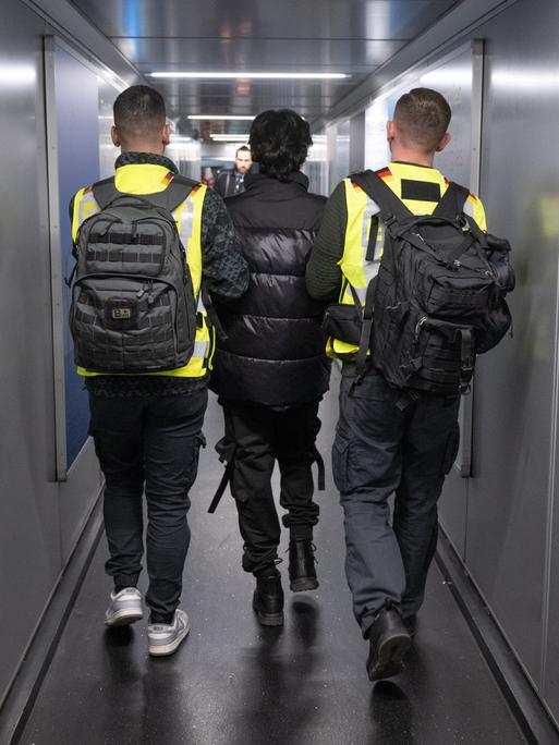 Zwei Beamte in Warnwesten eskortieren einen Mann durch einen Flugzeugtunnel.