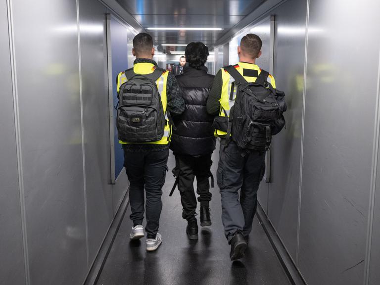 Zwei Beamte in Warnwesten eskortieren einen Mann durch einen Flugzeugtunnel.
