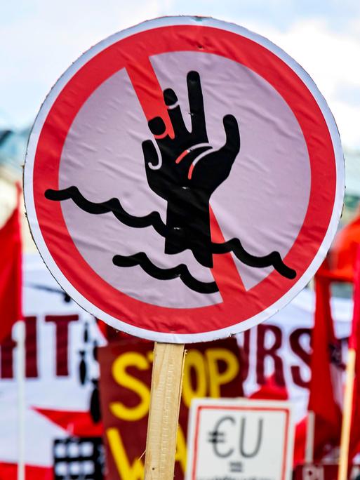 Ein rundes Schild mit der einem Piktogramm einer rot durchgestrichenen Hand in den Wellen versinkend, ist auf einer Demonstration in Stuttgart zu sehen.