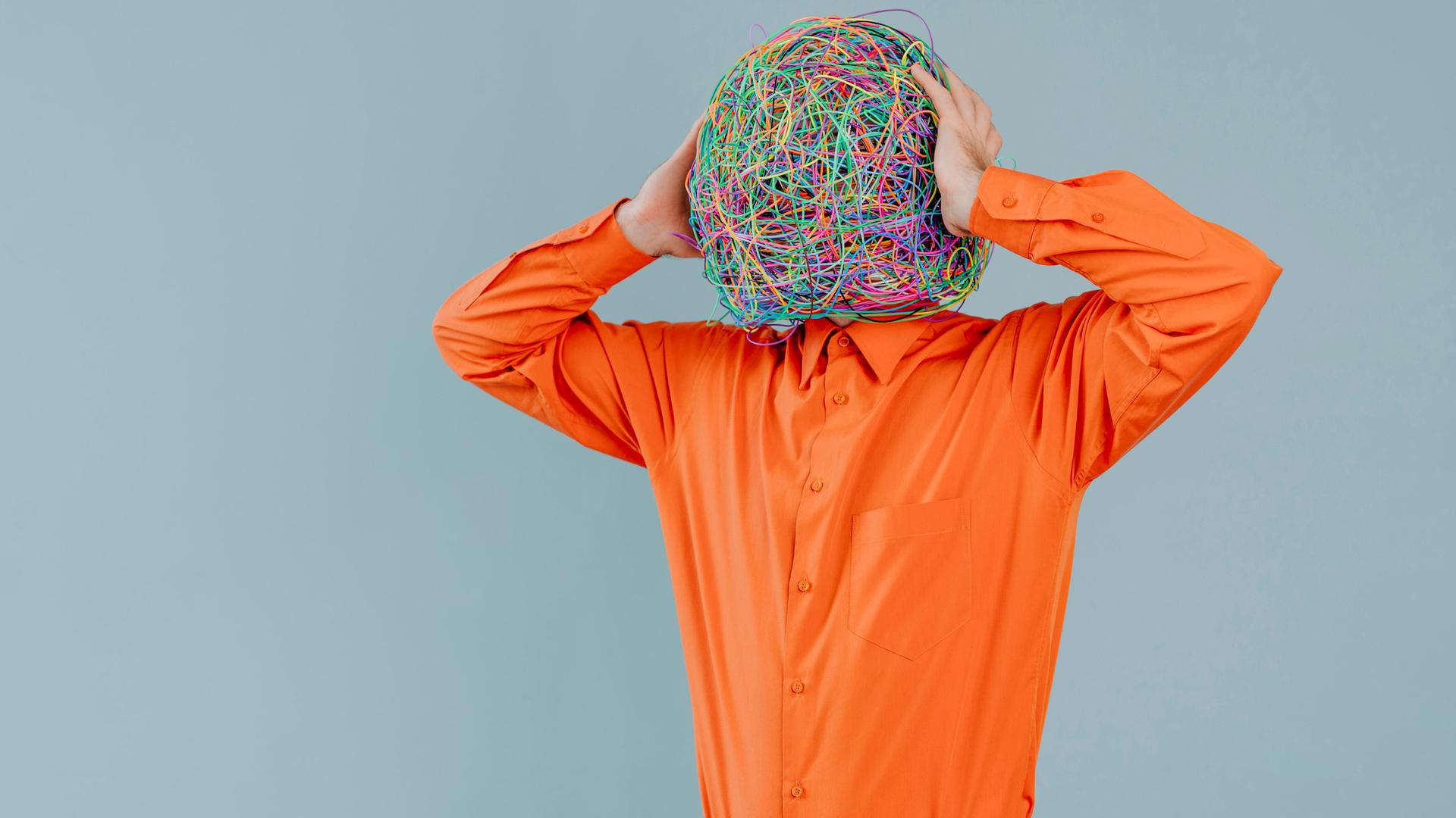 Ein Mann hält seine Hände an die Seiten seines Kopfs. Der Kopf ist in dem Bild eine große Kugel aus bunten Kabeln. Der Mann trägt ein orangefarbenes Hemd.