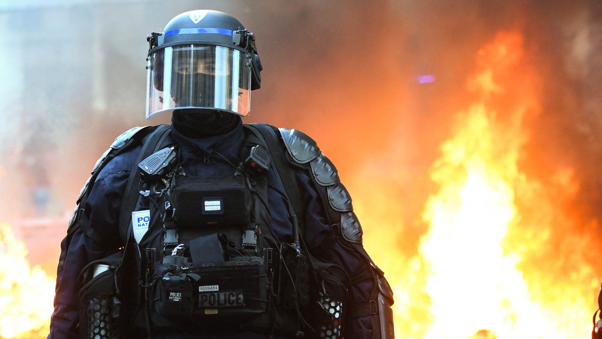Ein behelmter Polizist in voller Montur und Gesichtsmaske steht vor einem brennendem Hintergrund