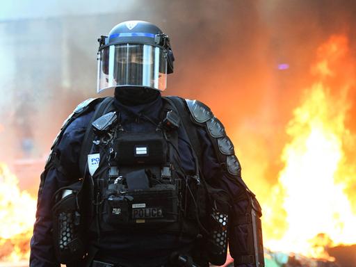 Ein behelmter Polizist in voller Montur und Gesichtsmaske steht vor einem brennendem Hintergrund Ein behelmter Polizist in voller Montur und Gesichtsmaske steht vor einem brennendem Hintergrund