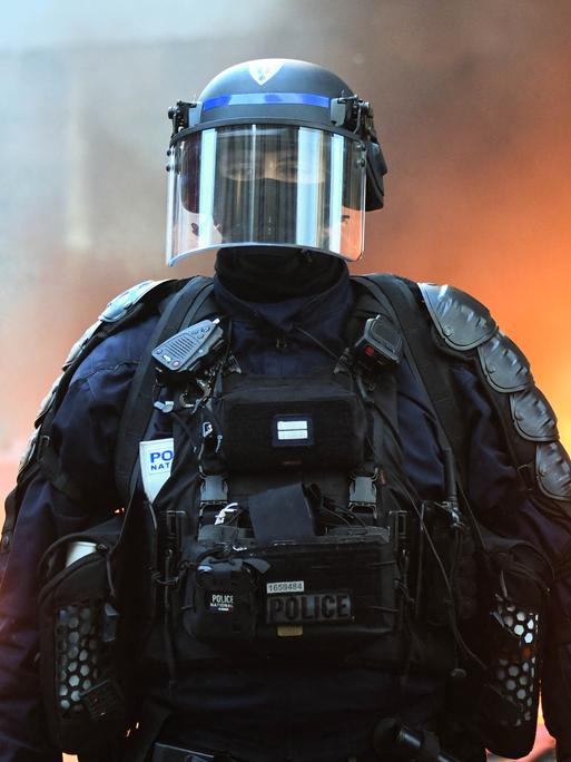 Ein behelmter Polizist in voller Montur und Gesichtsmaske steht vor einem brennendem Hintergrund Ein behelmter Polizist in voller Montur und Gesichtsmaske steht vor einem brennendem Hintergrund