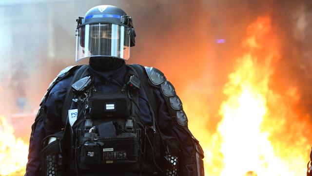 Ein behelmter Polizist in voller Montur und Gesichtsmaske steht vor einem brennendem Hintergrund