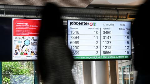 In einem Jobsender werden Wartenummern auf einem Bildschirm angezeigt. Im Vordergrund ist ein Mensch.