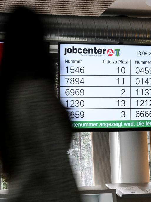 In einem Jobsender werden Wartenummern auf einem Bildschirm angezeigt. Im Vordergrund ist ein Mensch.