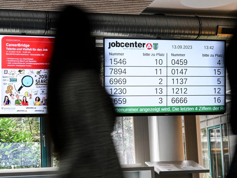 In einem Jobsender werden Wartenummern auf einem Bildschirm angezeigt. Im Vordergrund ist ein Mensch.