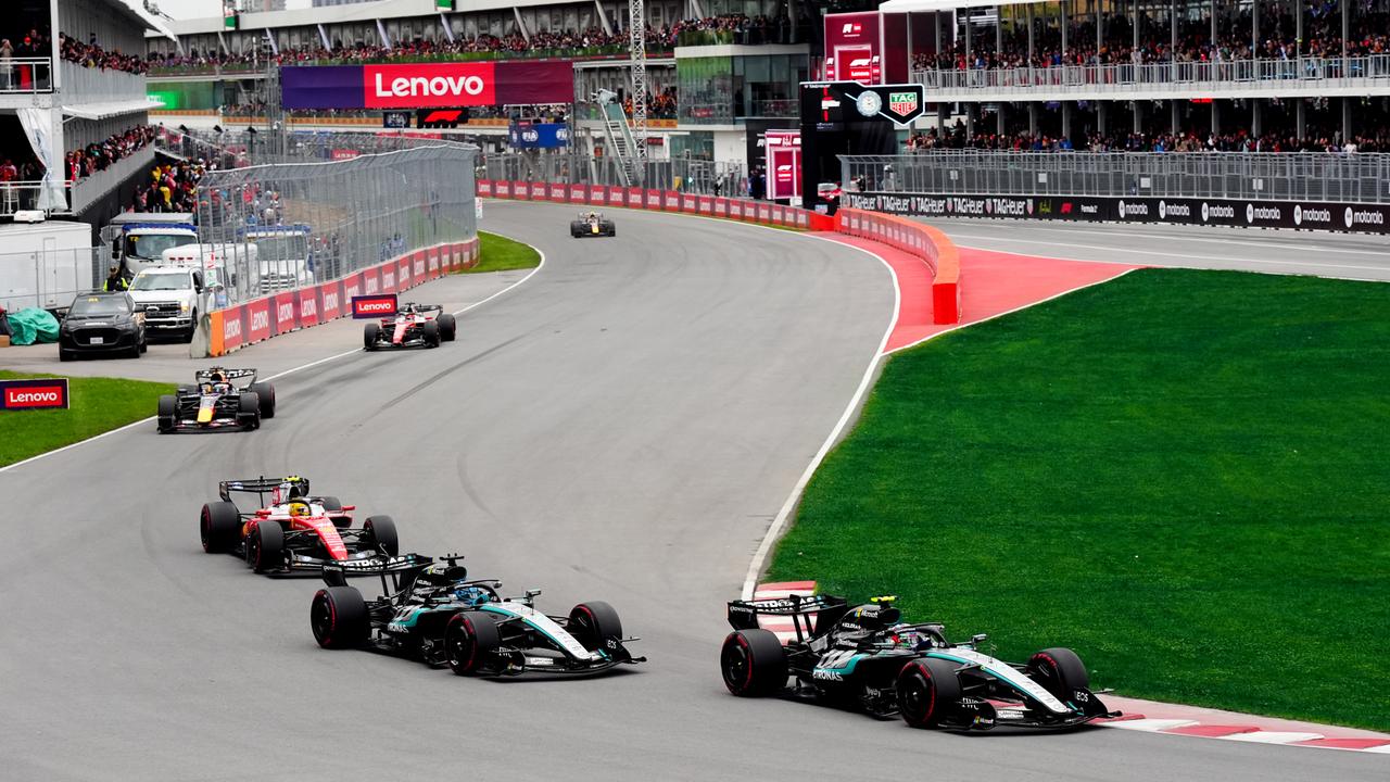 Zu sehen sind mehrere Boliden der Formel-1-Klasse auf einer Rennstrecke. Im Hintergrund sind Tribünen mit Zuschauern zu sehen.