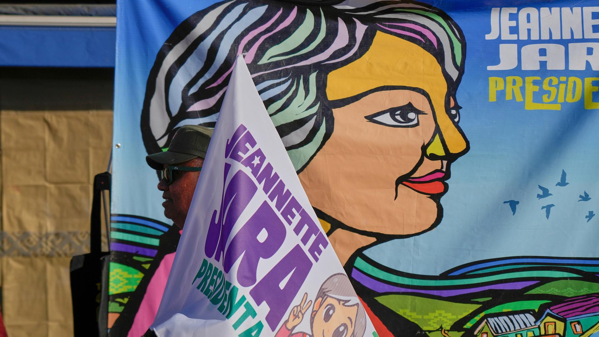 Vor einem großen Plakat mit einer stilisierten Darstellung Jaras sieht man den oberen Teil einer Fahne ebenfalls mit dem Namen und einer Darstellung Jaras.