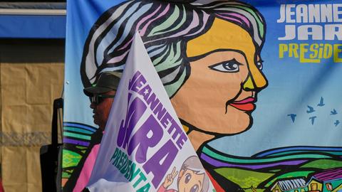 Vor einem großen Plakat mit einer stilisierten Darstellung Jaras sieht man den oberen Teil einer Fahne ebenfalls mit dem Namen und einer Darstellung Jaras.