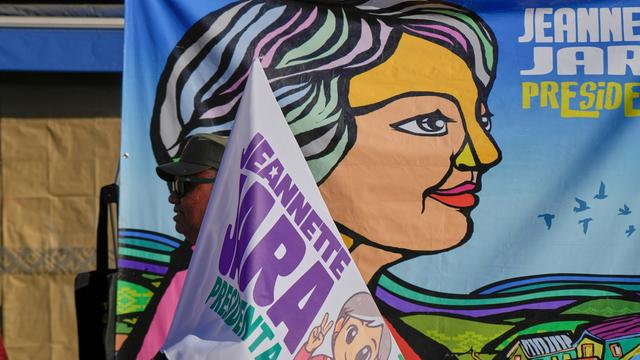 Vor einem großen Plakat mit einer stilisierten Darstellung Jaras sieht man den oberen Teil einer Fahne ebenfalls mit dem Namen und einer Darstellung Jaras.