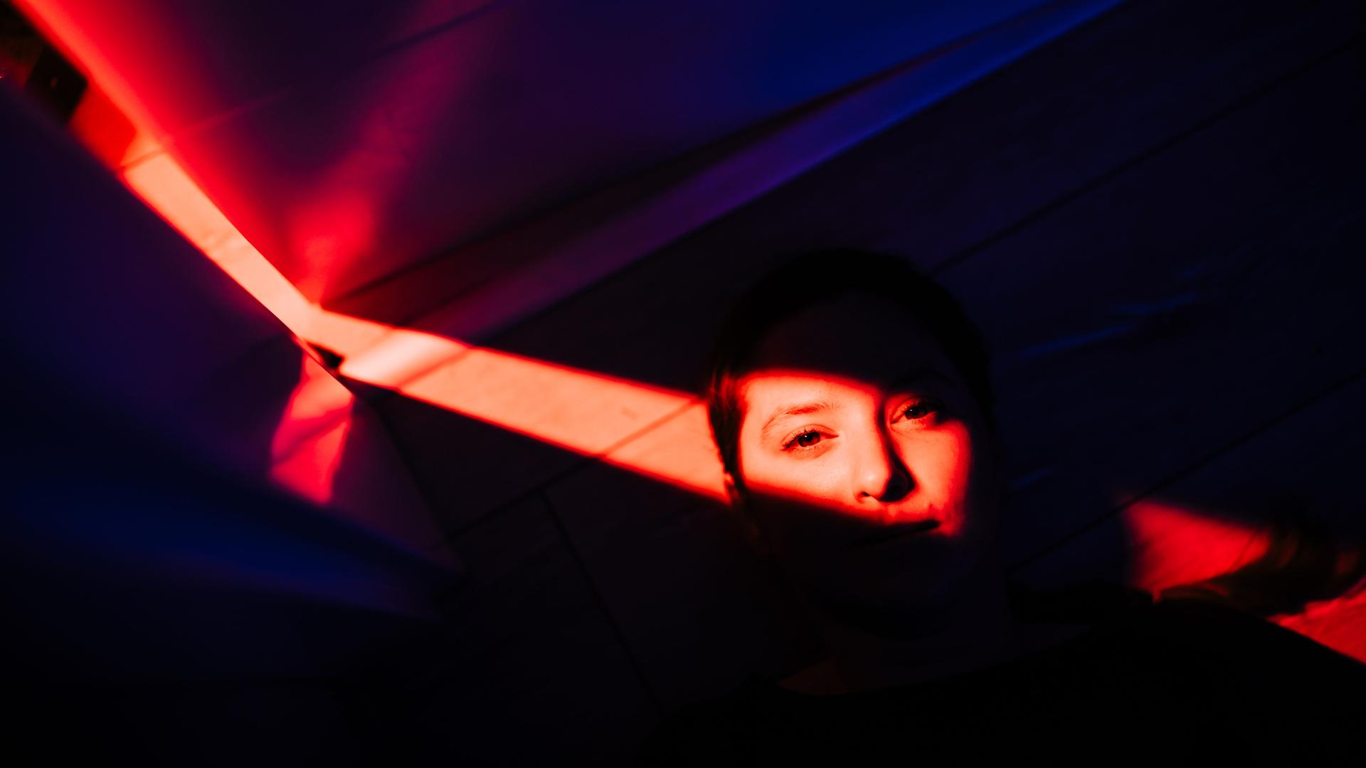 Porträt einer Frau, die auf dem Boden liegt. Ihr Gesicht ist nur fragmentarisch zu erkennen und wird von rotem Licht überdeckt. Der Rest des Bildes liegt im Schatten.