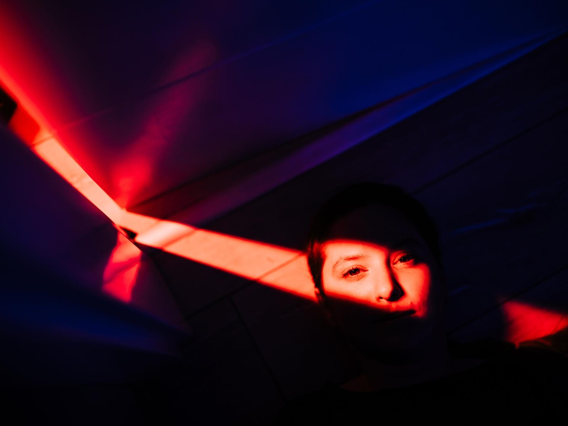 Porträt einer Frau, die auf dem Boden liegt. Ihr Gesicht ist nur fragmentarisch zu erkennen und wird von rotem Licht überdeckt. Der Rest des Bildes liegt im Schatten.