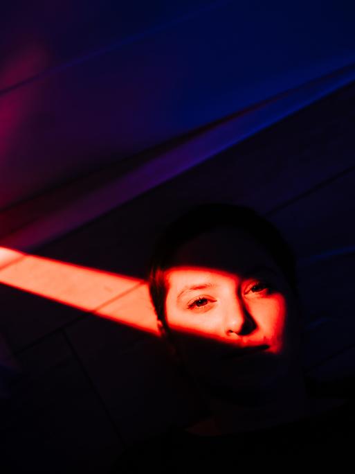 Porträt einer Frau, die auf dem Boden liegt. Ihr Gesicht ist nur fragmentarisch zu erkennen und wird von rotem Licht überdeckt. Der Rest des Bildes liegt im Schatten.