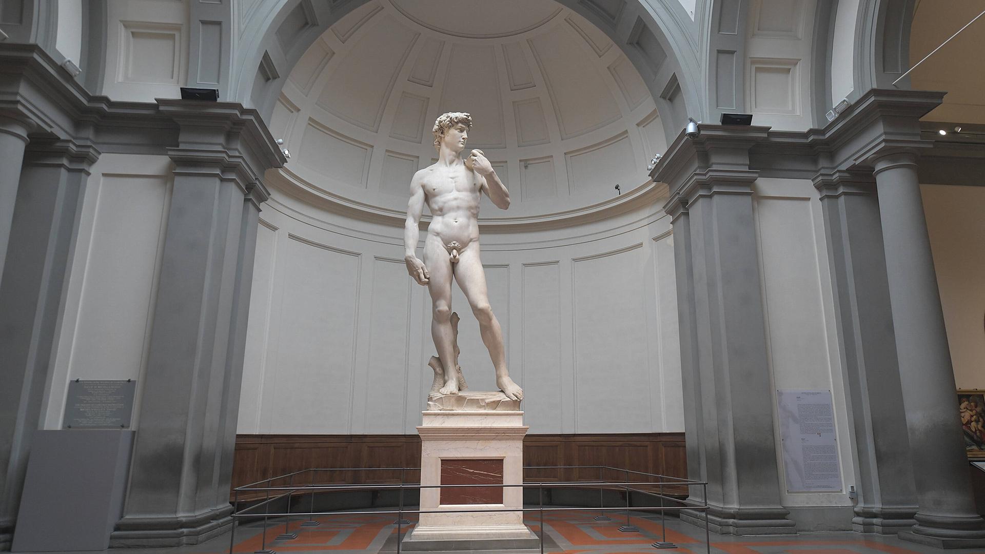 Die übergroße weiße Skulptur eines nackten Mannes steht in entspannter Pose auf einem Sockel allein in einem Museumsraum.