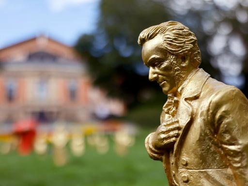 Eine goldene Richard-Wagner-Figuren stehen vor dem Festspielhaus. Der Komponist hat seine recht Hand auf die Brust gelegt und neigt sich leicht nach vorn.