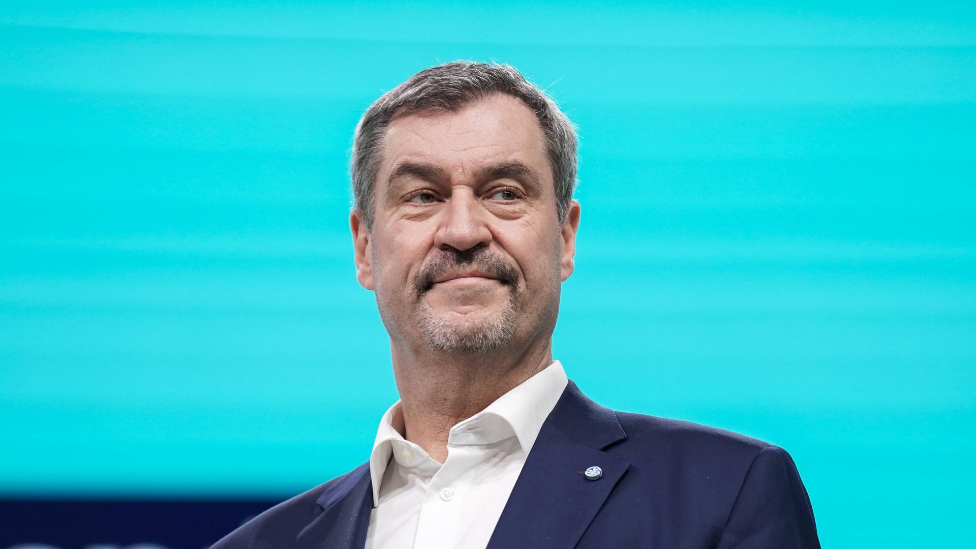 Markus Söder steht im dunklen Anzug auf einer Bühne vor einem leuchtend blauen Hintergrund.