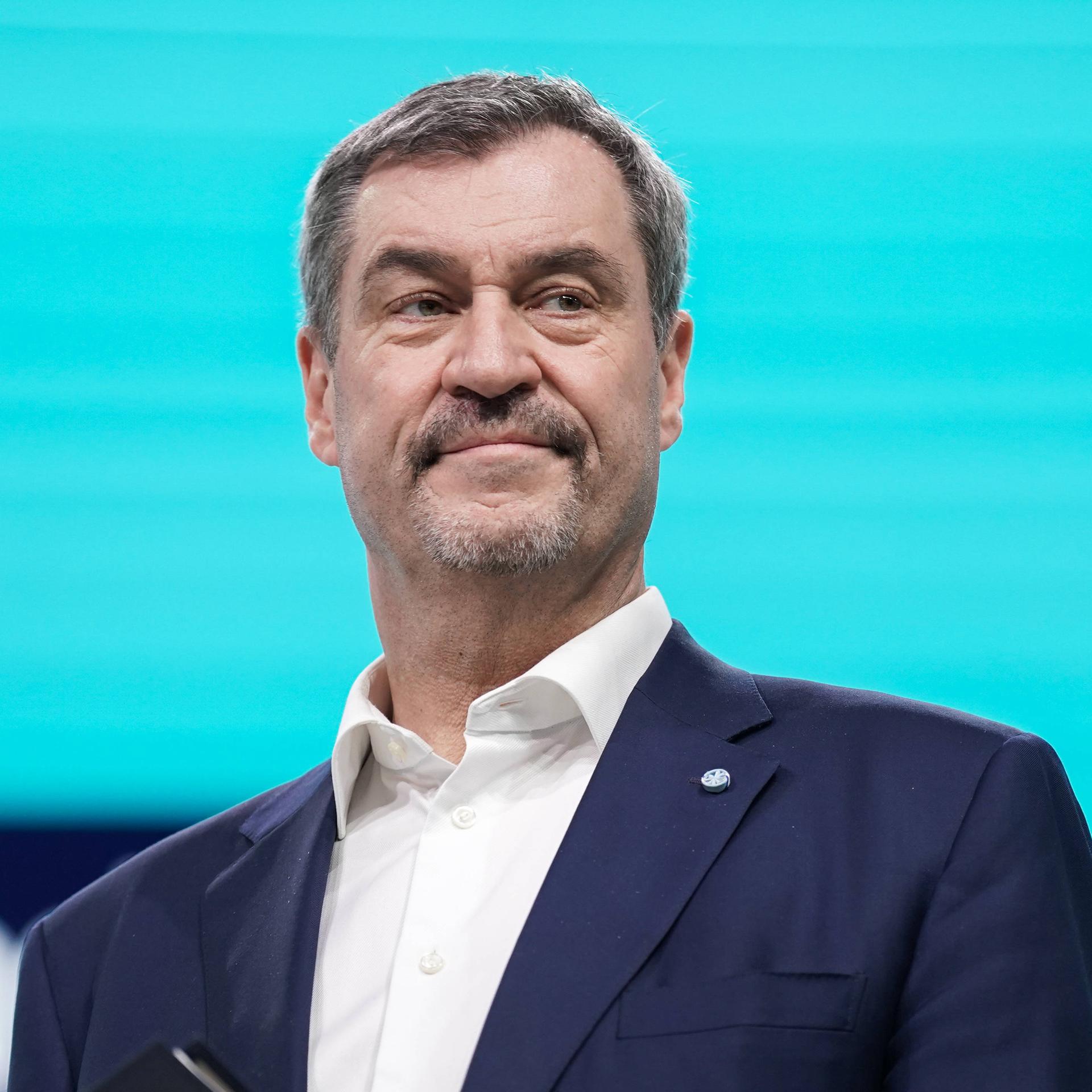 Markus Söder steht im dunklen Anzug auf einer Bühne vor einem leuchtend blauen Hintergrund.