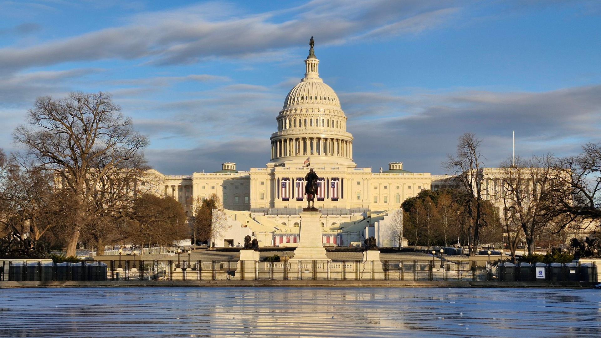 Blick auf das weiße Kapitol in Washington mit Kuppeldach. Im Vordergrund ein See und blätterlose Bäume.