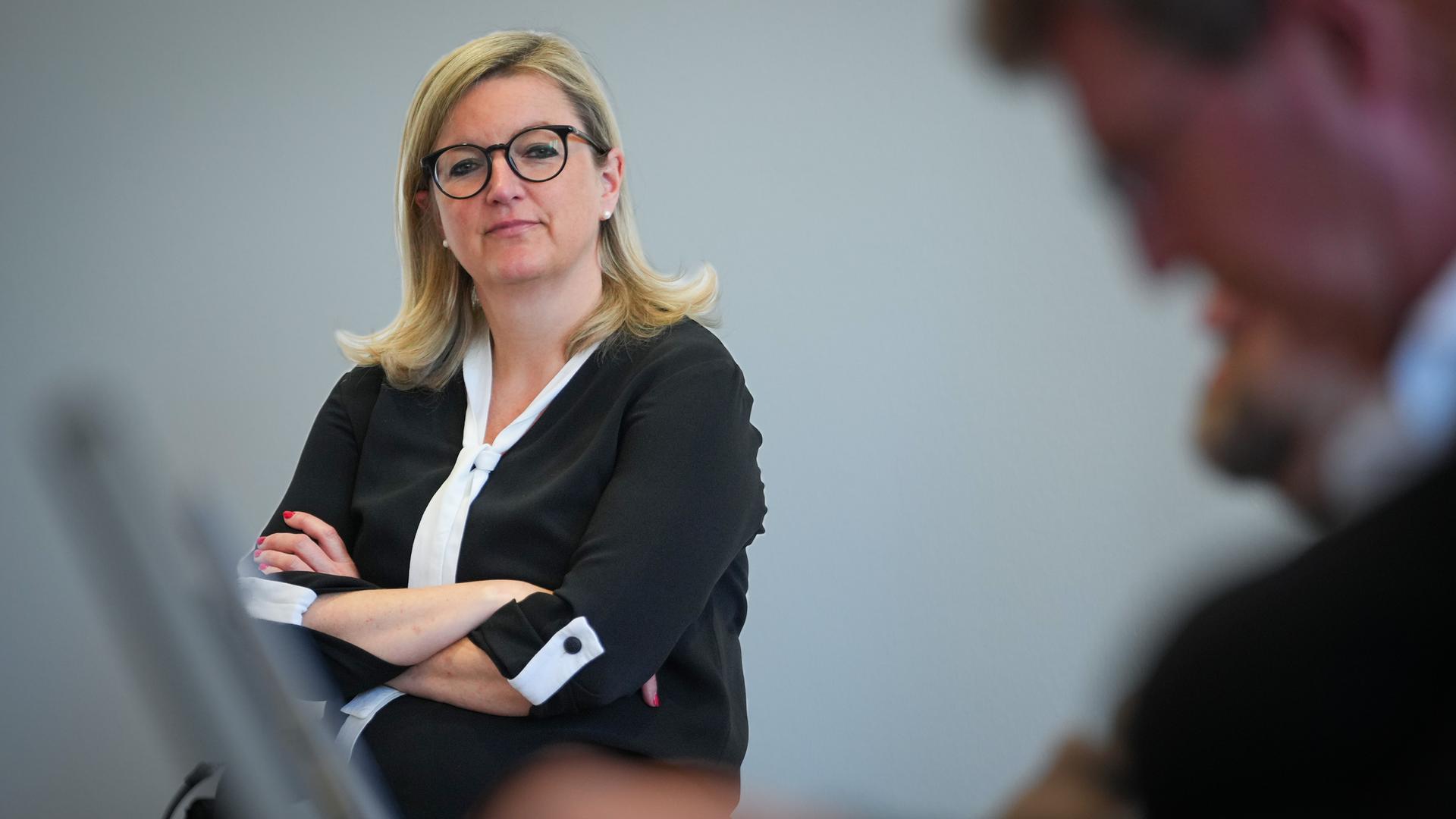 Brandenburgs Wirtschaftsministerin Klement hat die Arme verschränkt und den Kopf leicht schiefgelegt. Sie steht vor einem grauen Hintergrund.