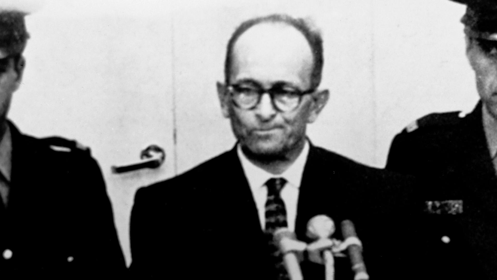 Adolf Eichmann steht in einer kugelsicheren Glaszelle im Gerichtssaal, umringt von Wachpersonal.