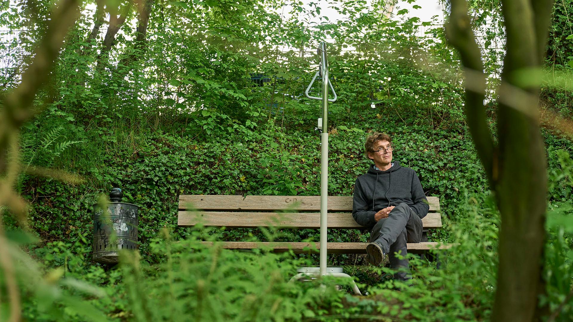 Ein Mann sitzt auf einer Bank in einem Park. Neben ihm steht ein graues Gestell, mit dem man sich im Krankenbett hochziehen kann. Er lauscht einer Klanginstallation.