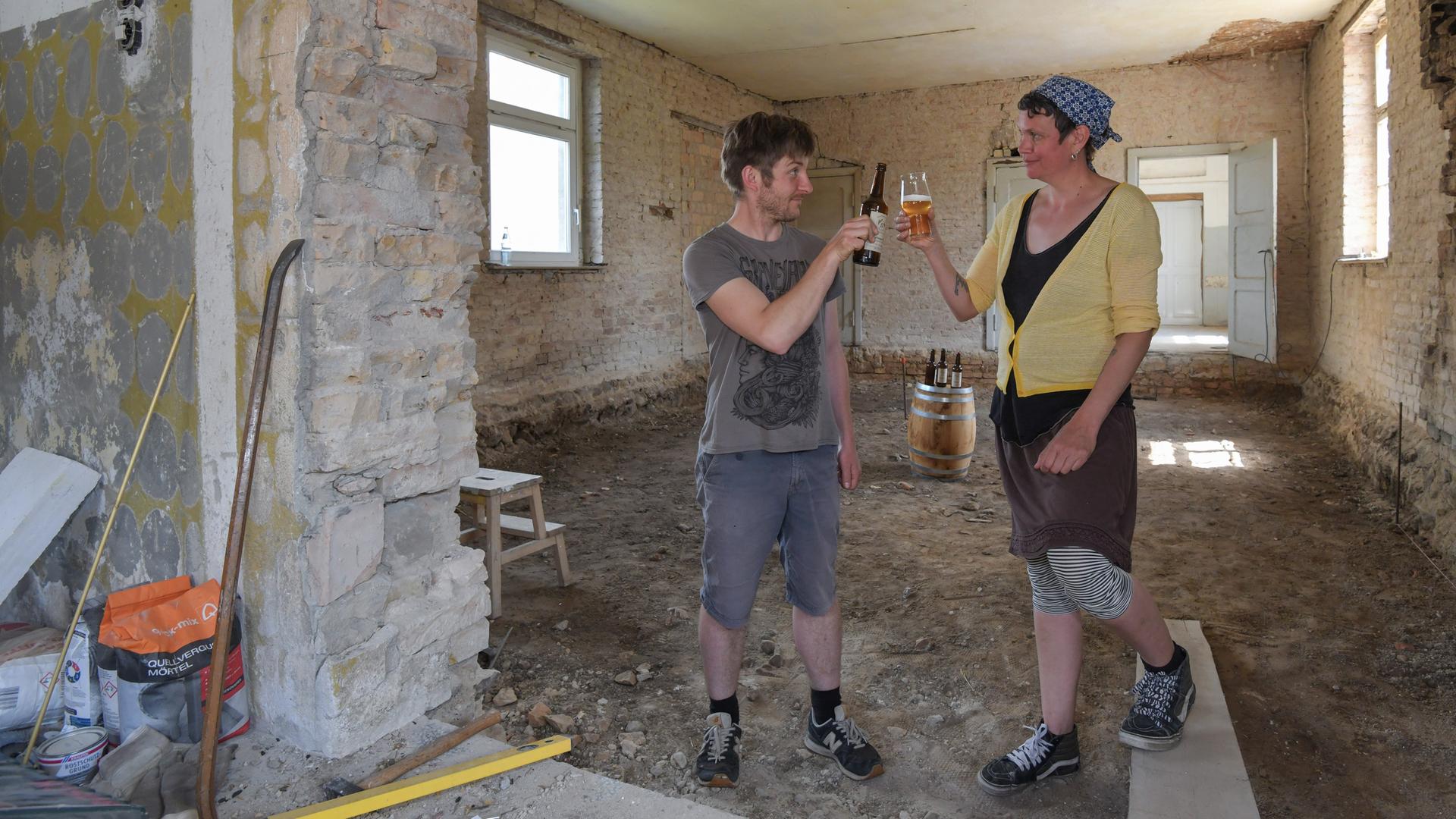 Zwei Personen stehen auf einer Baustelle in einem alten Klinkergebäude und stoßen mit Biergläsern an.