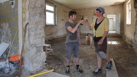 Zwei Personen stehen auf einer Baustelle in einem alten Klinkergebäude und stoßen mit Biergläsern an.