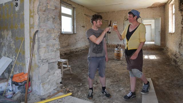 Zwei Personen stehen auf einer Baustelle in einem alten Klinkergebäude und stoßen mit Biergläsern an.