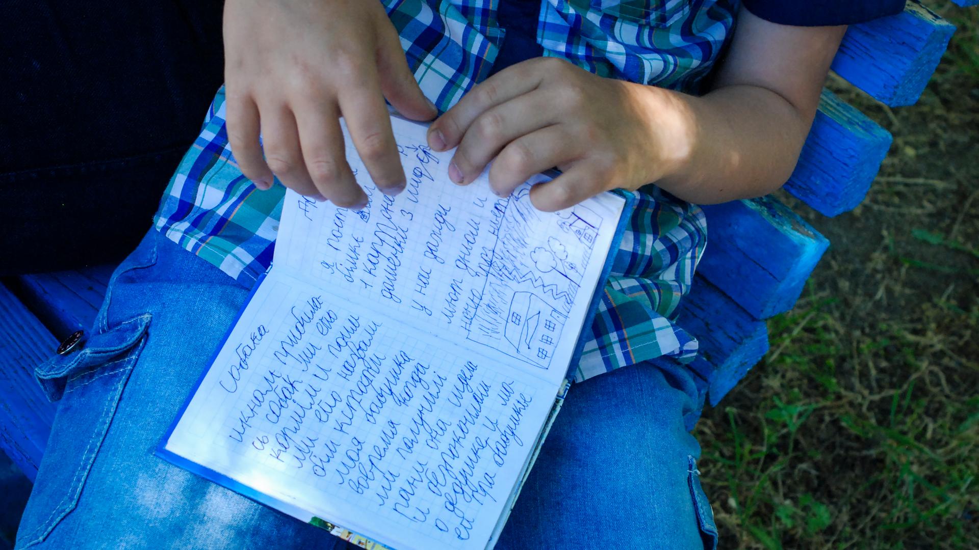 Der achtjährige Egor hält ein kleines blaues Buch in seiner Hand. Er schrieb in Mariupol Tagebuch, als um die Stadt blutig gekämpft wurde. Auf der aufgeschlagenen Seite steht neben einer kleinen Kinderzeichnung: "Ich habe gut geschlafen, habe gelächelt und 25 Seiten gelesen. Außerdem ist mein Großvater gestorben am 26.April. Saporischschja, 3. Juni 2022."