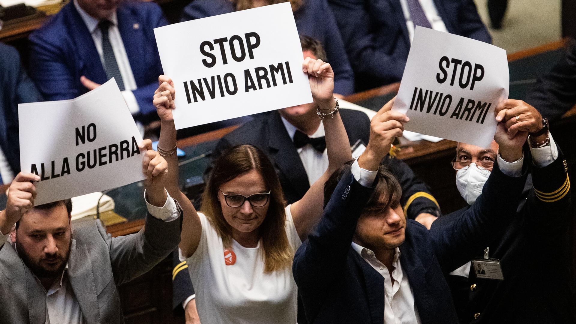 Im italienischen Parlament halten Abgeordnete der Opposition  - zwei Männer und eine Frau - weiße Plakate mit schwarzer Schrift hoch, die sich inhaltlich gegen Waffenlieferungen in die Ukraine und gegen den Krieg richten: "No Alla Guerra" und "Stop Invio Armi" lauten die Schriftzüge auf den Plakaten.