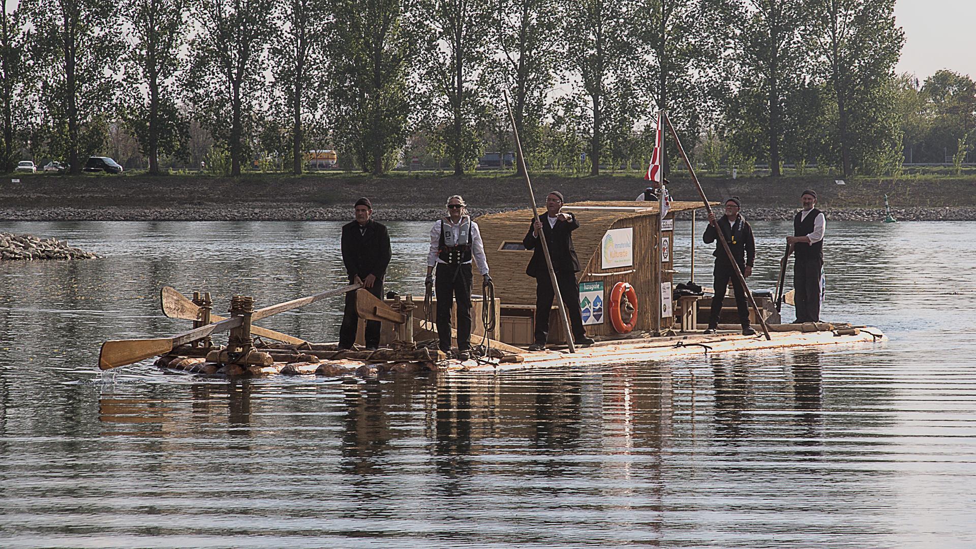 Fünf Männer stehen auf einem Holzfloß, der im Fluss treibt.