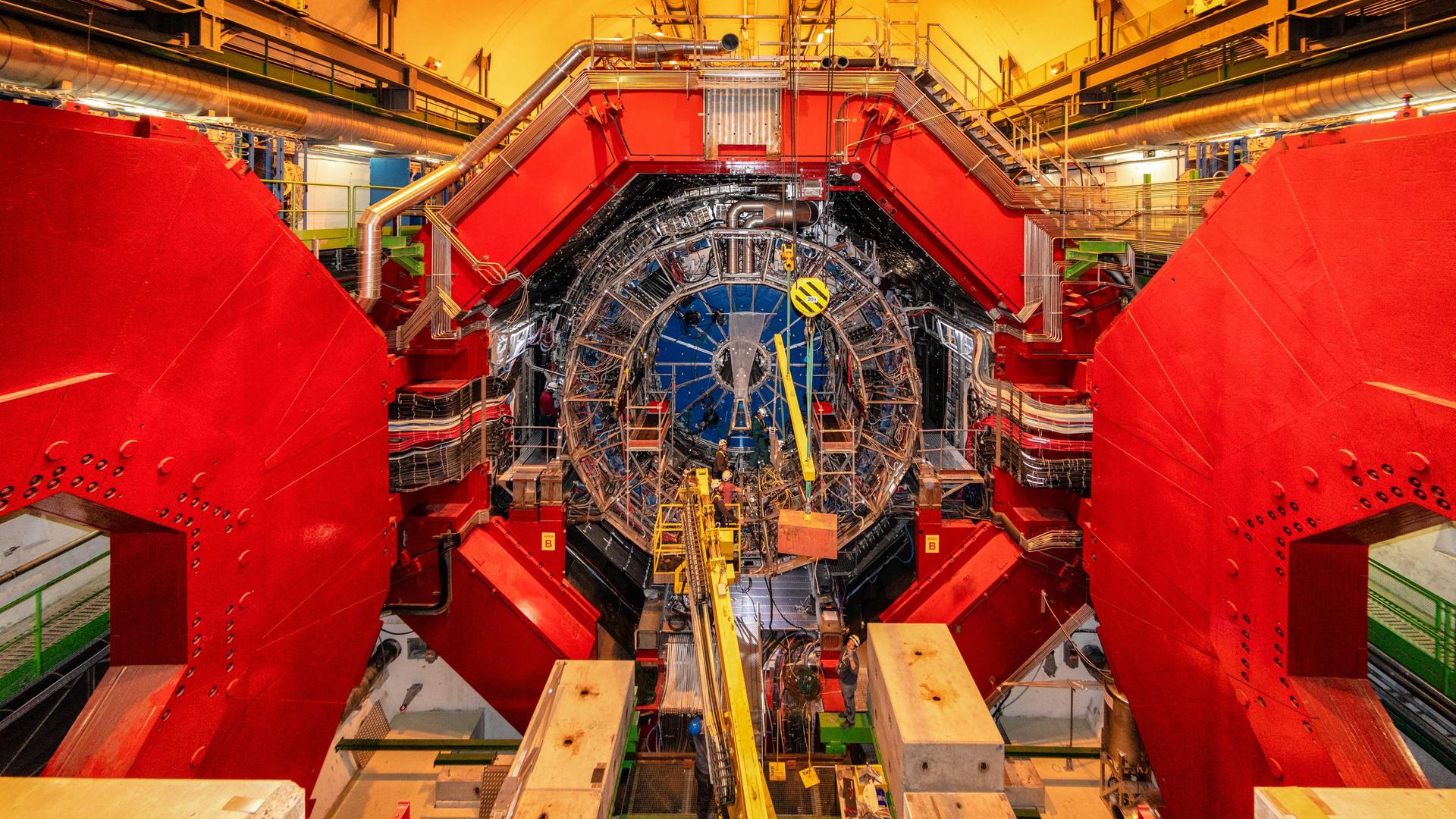 Blick in eine Halle der Europäischen Organisation für Kernforschung. Zu sehen ist ein überdimensional großer Schwerionendetektor auf dem zwei Personen stehen, die nur circa ein Zehntel so groß sind wie der Detektor an sich.
