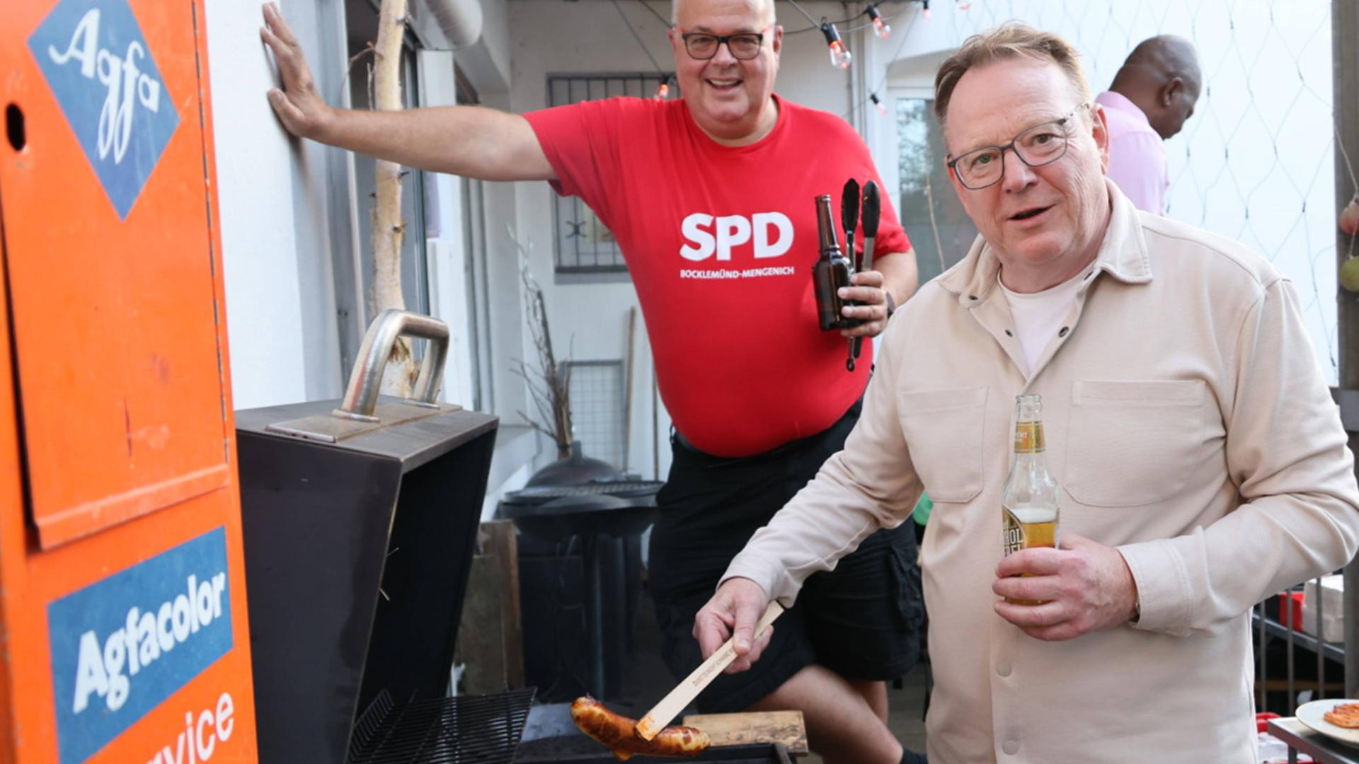 Torsten Burmester steht an einem Grill und hält mit einer Zange eine Bratwurst. Im Hintergrund steht ein Mann in rotem SPD-Shirt mit einem Bier in der Hand. Torsten Burmester steht an einem Grill und hält mit einer Zange eine Bratwurst. Im Hintergrund steht ein Mann in rotem SPD-Shirt mit einem Bier in der Hand.