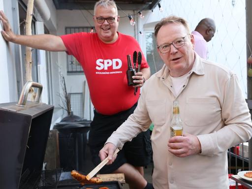 Torsten Burmester steht an einem Grill und hält mit einer Zange eine Bratwurst. Im Hintergrund steht ein Mann in rotem SPD-Shirt mit einem Bier in der Hand.