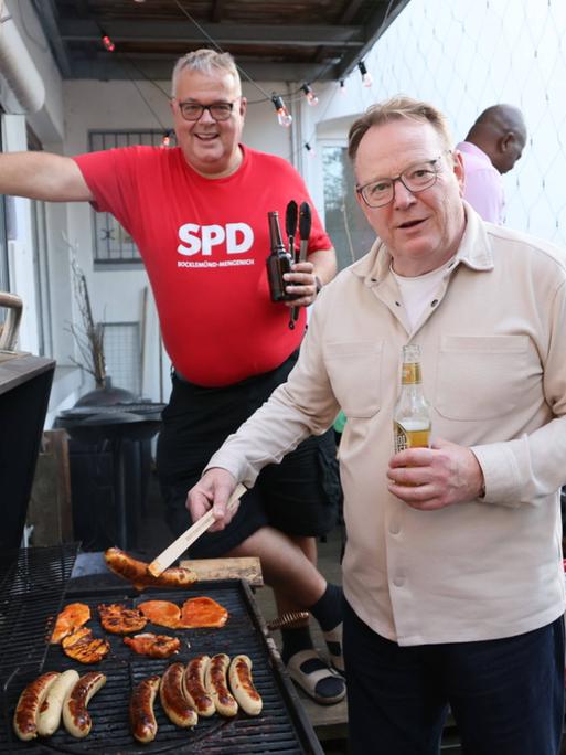 Torsten Burmester steht an einem Grill und hält mit einer Zange eine Bratwurst. Im Hintergrund steht ein Mann in rotem SPD-Shirt mit einem Bier in der Hand.