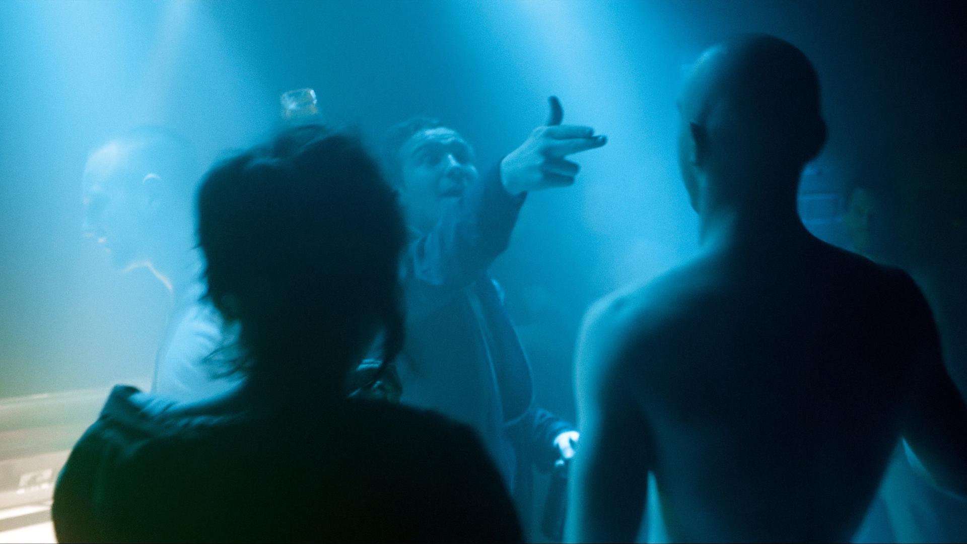 Laia Costa, Frederick Lau und Franz Rogowski in Sebastian Schippers "Victoria". Frederick Lau richtet eine imaginäre Pistole auf die anderen, alle stehen in blauem Club-Dämmerlicht. 