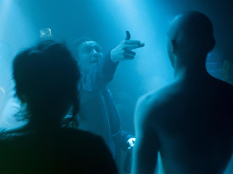 Laia Costa, Frederick Lau und Franz Rogowski in Sebastian Schippers "Victoria". Frederick Lau richtet eine imaginäre Pistole auf die anderen, alle stehen in blauem Club-Dämmerlicht. 