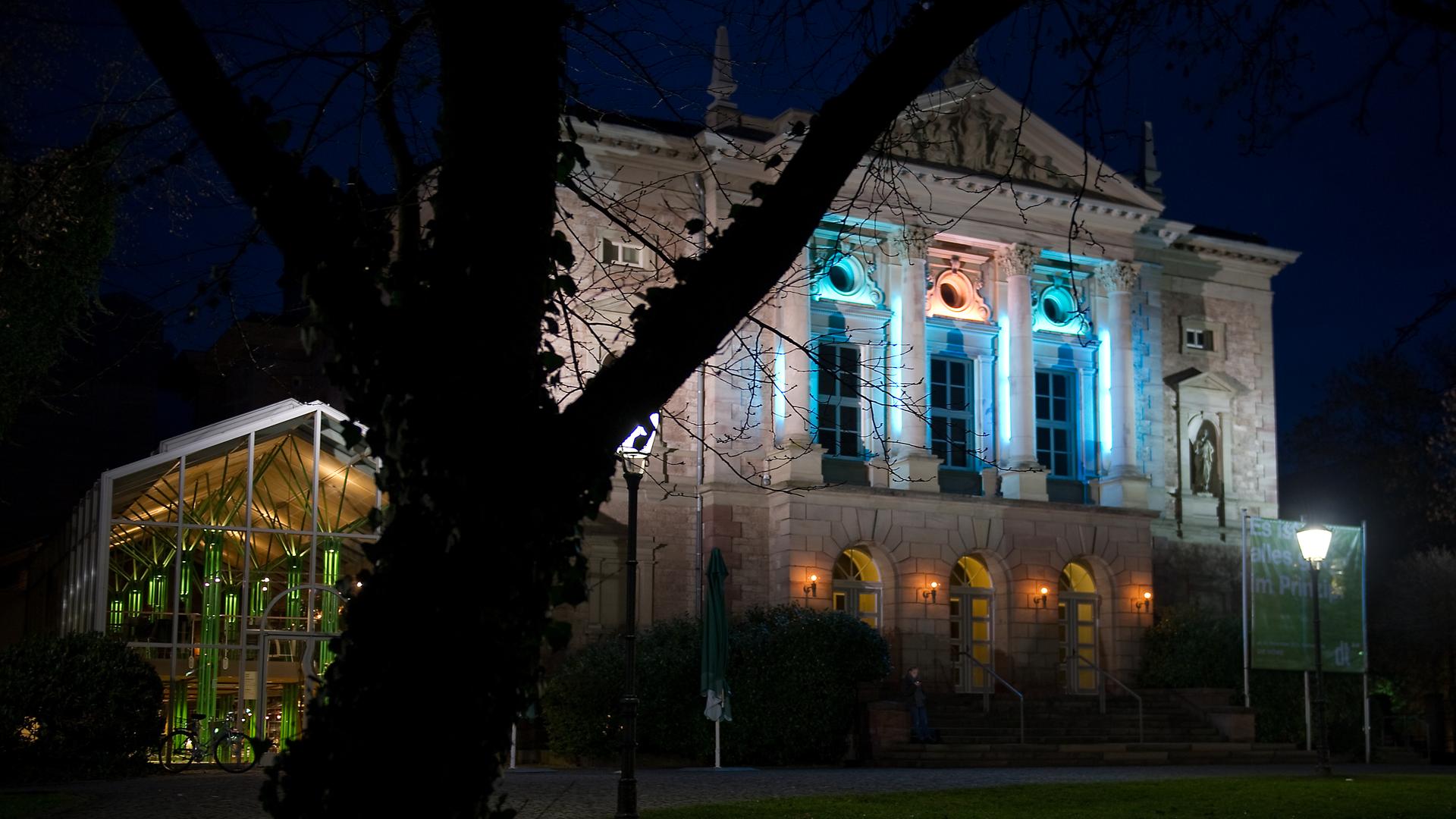 Das Bild zeigt das Deutsche Theater in Göttingen nachts mit beleuchteten Fenstern. Es ist ein altes Gebäude mit hohen Fenstern und Stuckverzierungen an der Fassade. 
