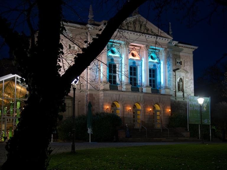 Das Bild zeigt das Deutsche Theater in Göttingen nachts mit beleuchteten Fenstern. Es ist ein altes Gebäude mit hohen Fenstern und Stuckverzierungen an der Fassade. 
