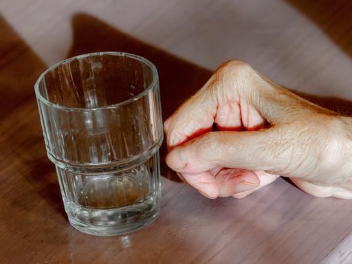 Die Hand einer alten Frau liegt neben einem leeren Wasserglas auf einem Tisch.