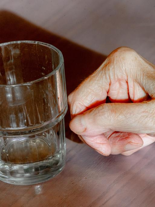 Die Hand einer alten Frau liegt neben einem leeren Wasserglas auf einem Tisch.