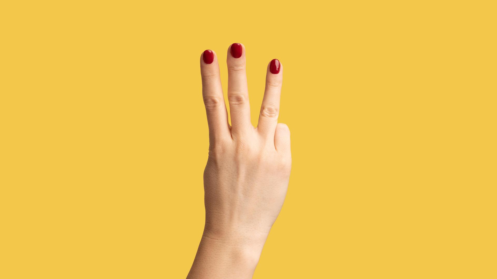 Eine schlanke Hand mit rot lackierten Fingenägeln zeigt vor gelbem Hintergrund drei Finger. Eine schlanke Hand mit rot lackierten Fingenägeln zeigt vor gelbem Hintergrund drei Finger.
