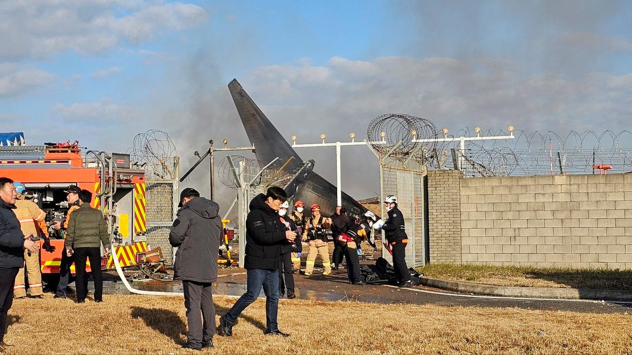 Man sieht die Heckflosse des Flugzeugs hinter einer Mauer. Es laufen Feuerwehrleute und andere Personen herum, links steht ein Feuerwehrauto. Rauch steigt auf.