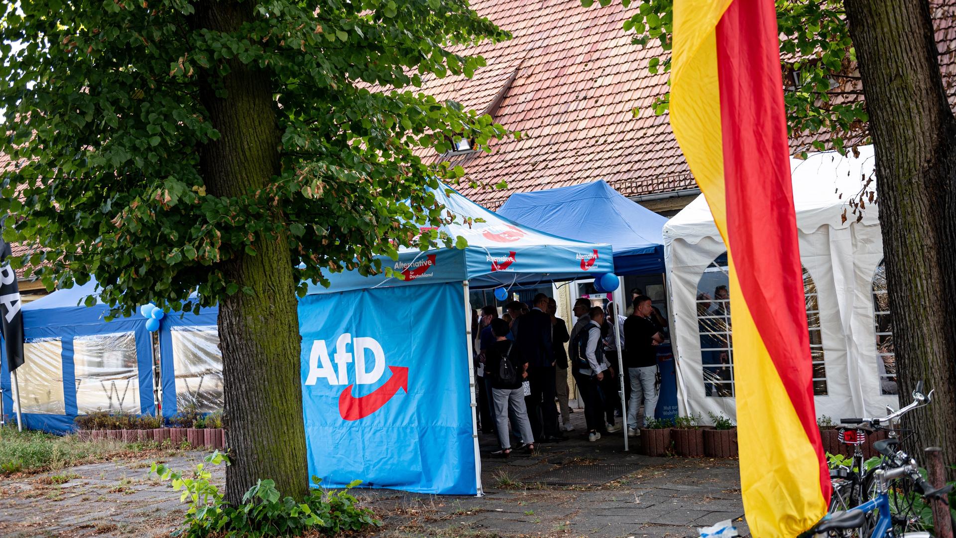 Unterstützer der AfD stehen bei einer Wahlparty der AfD in einem blauen Zeltpavillon mit dem Logo der Partei, davor weht eine Deutschlandflagge.