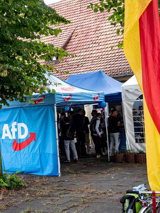 Unterstützer der AfD stehen bei einer Wahlparty der AfD in einem blauen Zeltpavillon mit dem Logo der Partei, davor weht eine Deutschlandflagge.