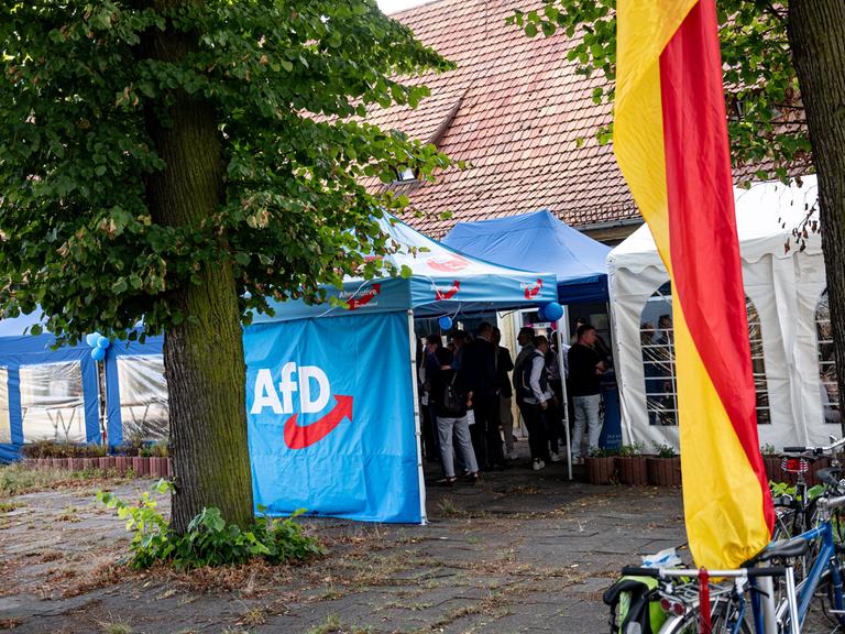 Unterstützer der AfD stehen bei einer Wahlparty der AfD in einem blauen Zeltpavillon mit dem Logo der Partei, davor weht eine Deutschlandflagge.