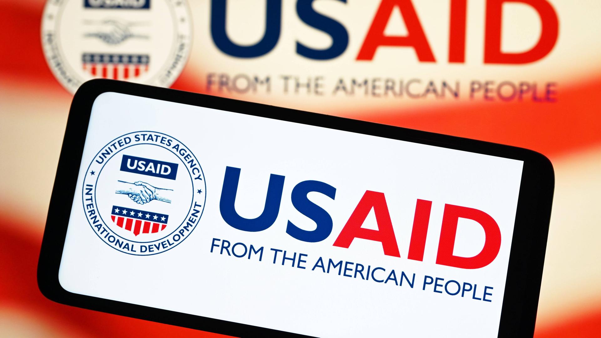 Das Land USA hat eine Behörde, sie sich um Entwicklungs-Hilfe für ärmere Länder kümmert. Die Behörde heißt USAID. Das Foto zeigt das Zeichen der Behörde USAID auf dem Bildschirm eines Handys.