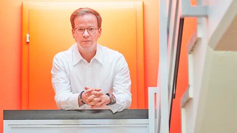 Ein schlanker, weißer Mann mit dunkelblonden Haaren und Brille posiert fürs Foto in einem Treppenhaus, die Wand und Tür hinter ihm ist leuchtend orange. Der Mann trägt ein weißes Hemd, ein Armband und eine auffällige Armbanduhr.